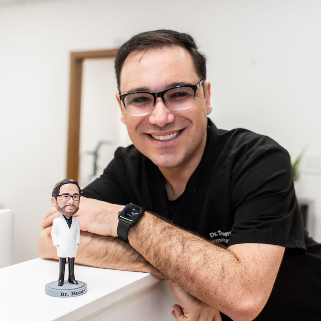 Dr. Danny Del Campo smiling in black scrubs next to a figurine of himself in a medical coat, representing Chicago Skin Clinic's expert dermatology services.